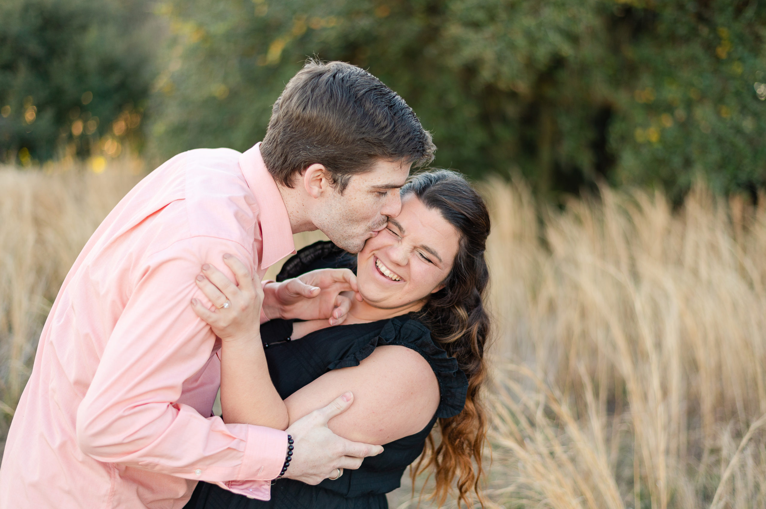 Engagement photo of a man pulling a woman close and kissing her on the temple while she is giggling