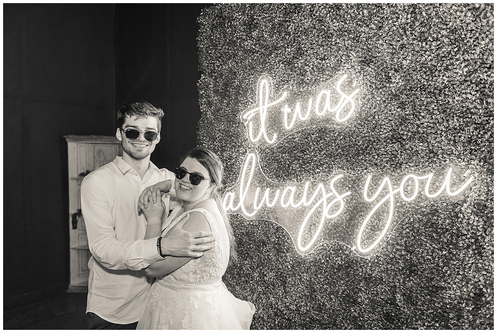 Bride and groom standing beside an acrylic neon sign that says "It was always you" while wearing sunglasses and smiling at the camera. 