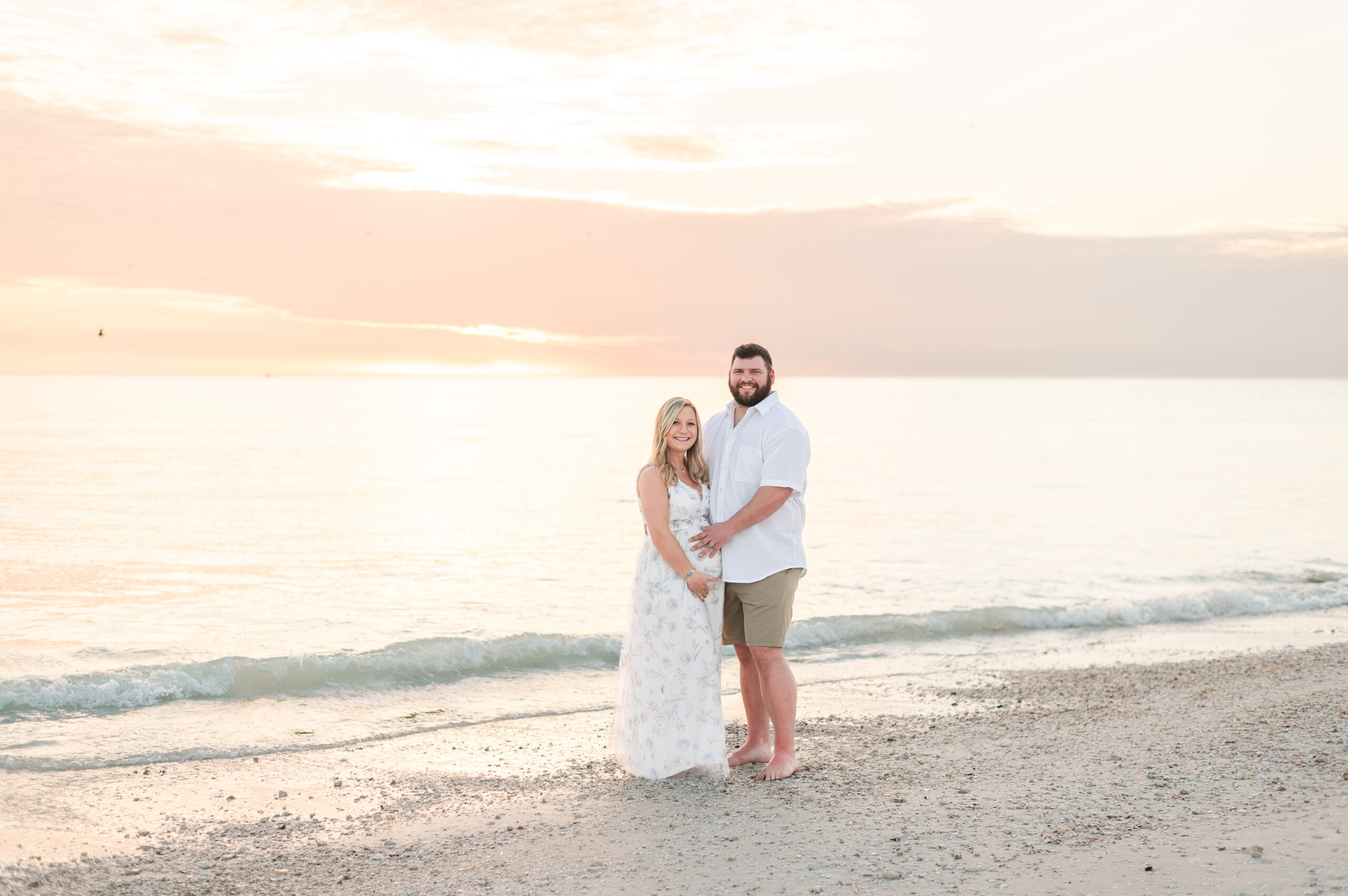 Couple standing at the waters edge at the beach with with beautiful sunset behind them. The woman is pregnant, and holding her belly and the husband is holding her belly too. She is wearing a long white dress and he is wearing a white shirt and khaki shorts.