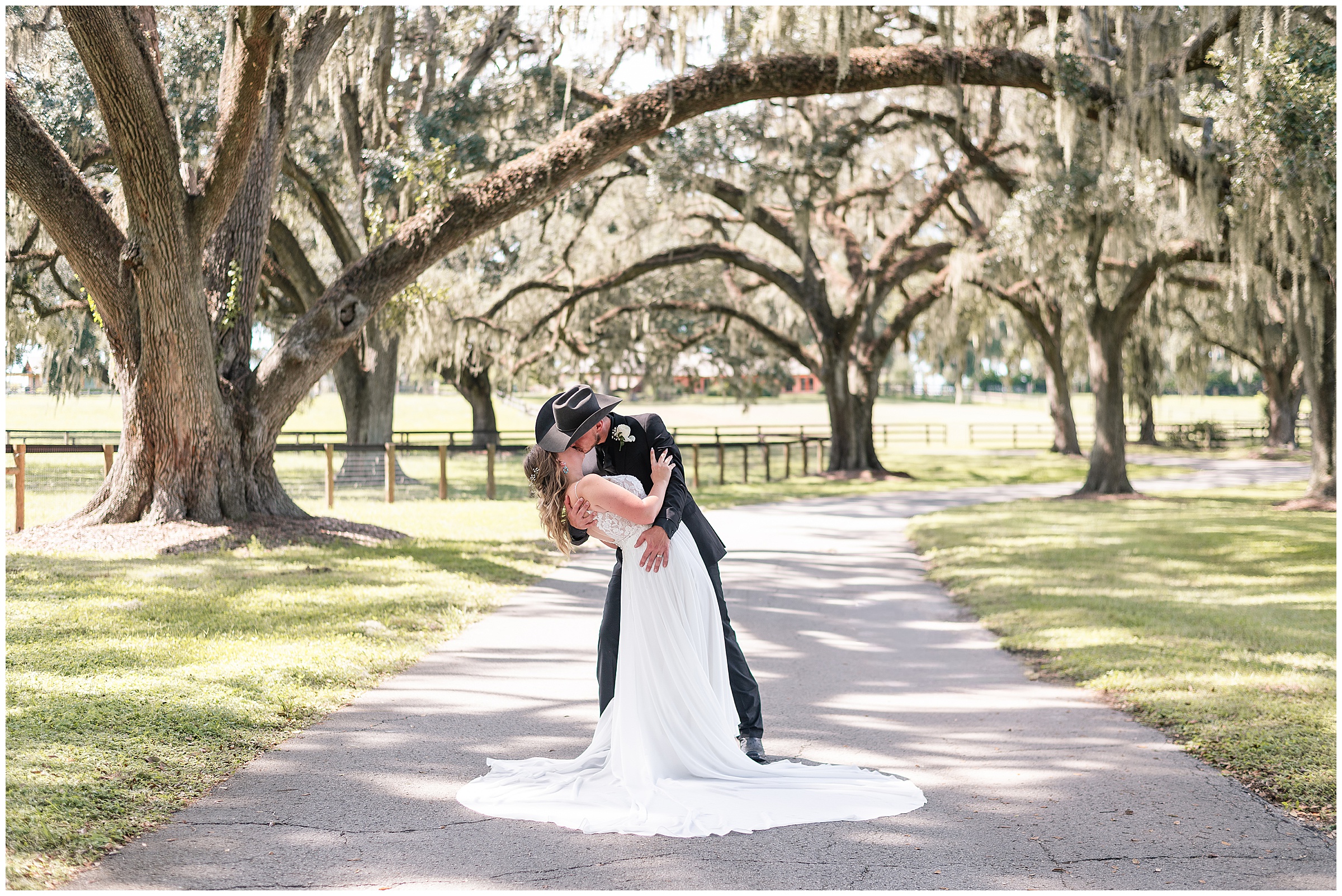 A groom dipping and kissing his bride while standing in the middle of the driveway underneath oak trees at their Ocala Wedding at Protea Weddings and Events. 