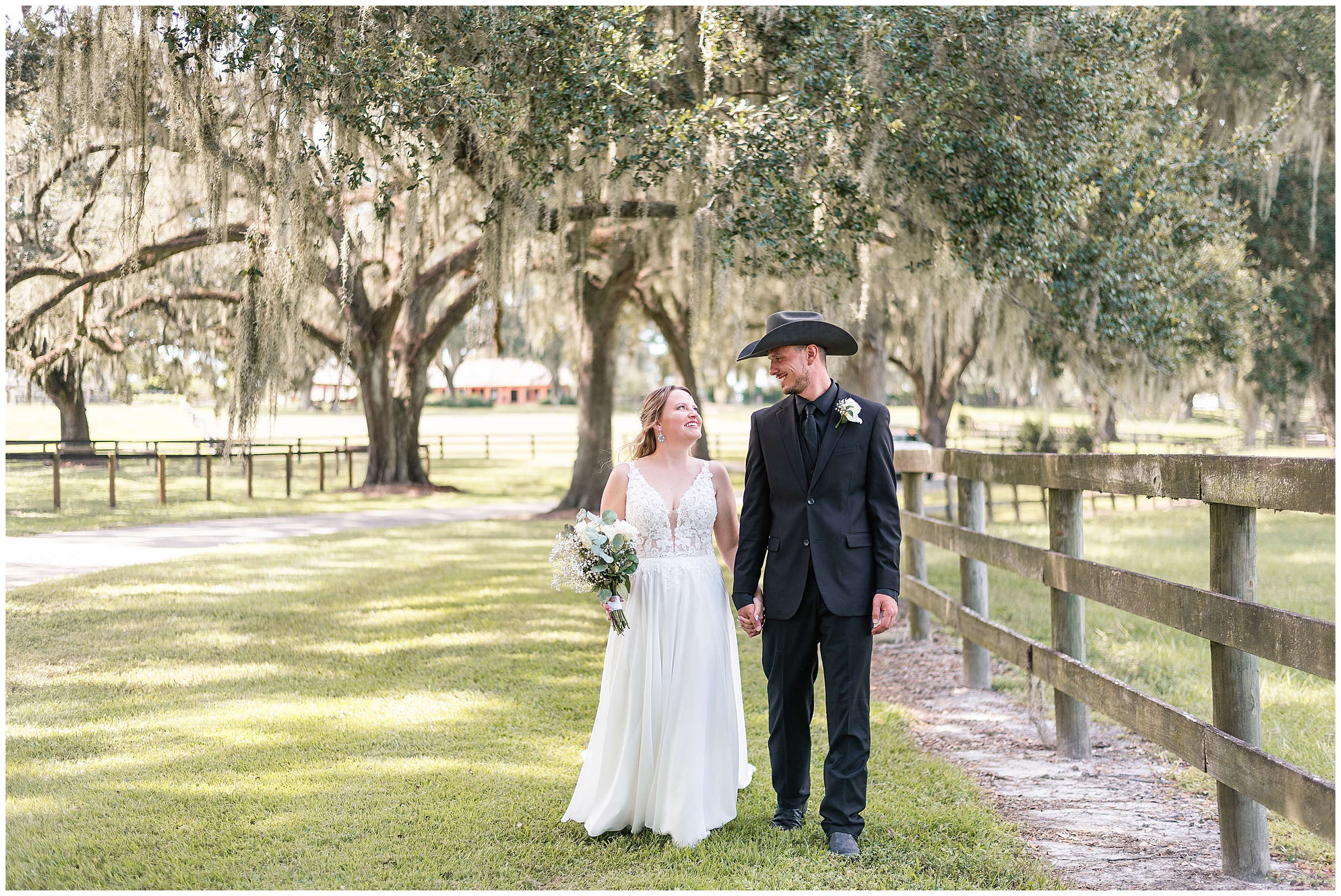 Bride and groom walking together along a fenceline at a farm with oak trees at their Ocala Wedding at Protea Weddings and Events. 